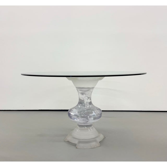 A sculptural dining table featuring a round glass top supported by an urn-shaped Lucite pedestal. The base combines a...