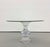 A sculptural dining table featuring a round glass top supported by an urn-shaped Lucite pedestal. The base combines a...