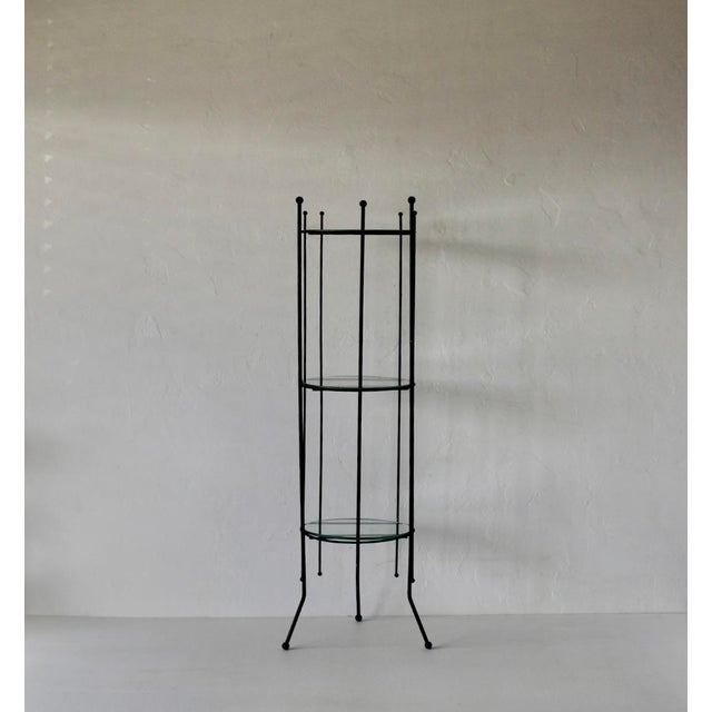 A narrow etagere featuring a matte black metal frame with glass shelves. Circular metal frame with three vertical supports...