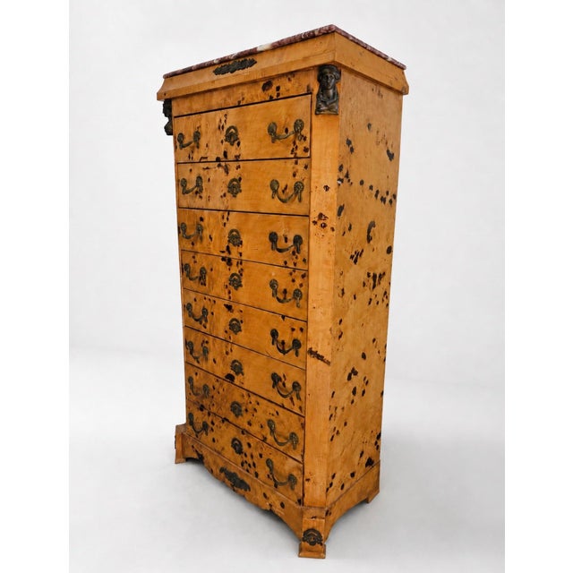 Tall chest of drawers with a warm burl wood finish, marble top, and decorative dark metal hardware. The piece has an...