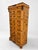 Tall chest of drawers with a warm burl wood finish, marble top, and decorative dark metal hardware. The piece has an...