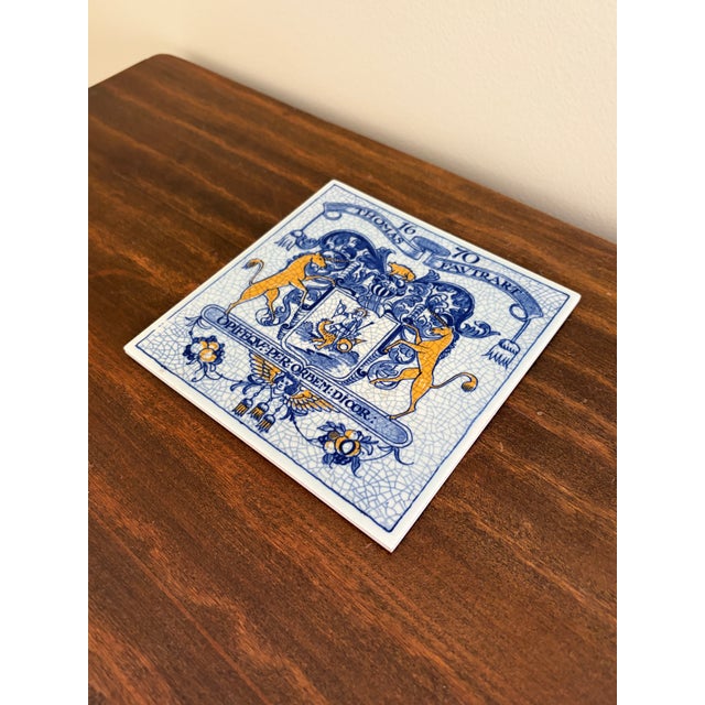 A classic Dutch Delft-style hand painted blue and white porcelain tile featuring an intricately rendered heraldic coat of...