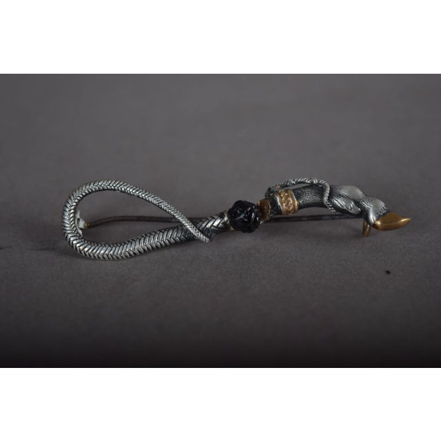 1930s brooch with the combined motif of a looping snake tail and a bull leg, in silver with gold-tone accents and a dark...