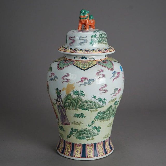 A large Chinese covered urn offers porcelain construction with figural foo dog finial over vessel with hand enameled genre...