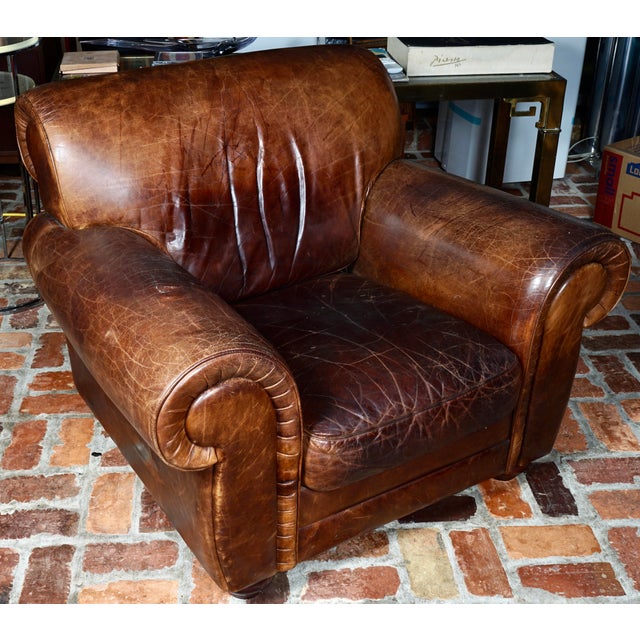 A vintage brown leather club chair featuring rolled arms and noticeable natural distressing and creasing on the leather....