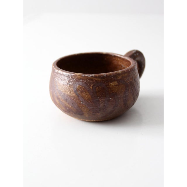 A vintage handmade studio pottery mug with a rounded, low form and a softly pulled handle. The surface is finished in a...