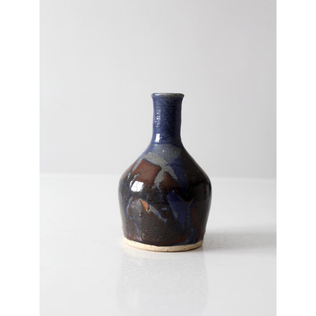 A vintage 1970s handmade studio pottery bud vase with a rounded bottle form and flowing blue and earthy brown glazes. The...