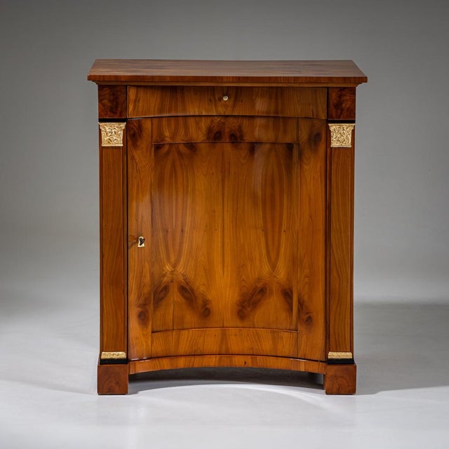 Biedermeier demi-cabinet in cherrywood with a concave front and flanking pilasters featuring gold-patinated capitals and...