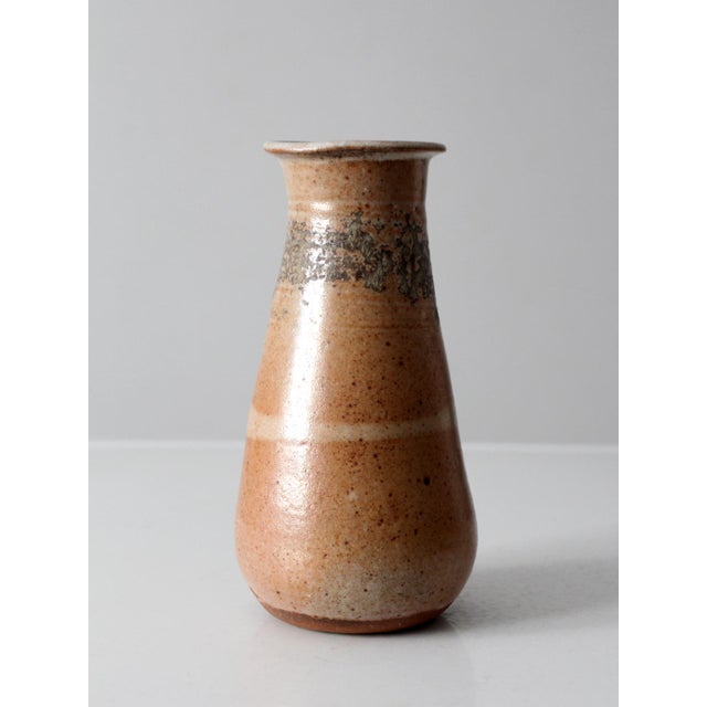 A vintage handmade studio pottery vase featuring a softly tapered form and a layered neutral glaze in warm peach and stone...