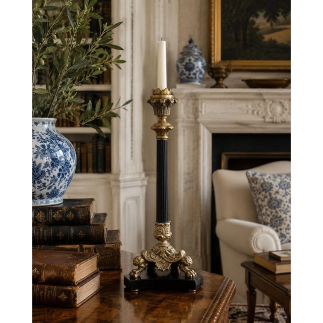 A tall neoclassical candlestick combining warm brass ornament with a slender, fluted black stem. The architectural base is...