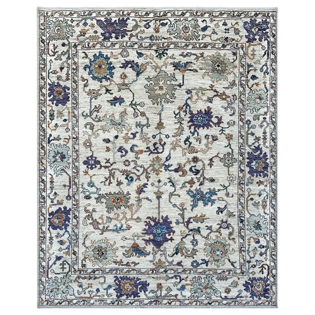 A light, airy ivory Oushak area rug, its creamy field drifting with deep violet and cornflower blossoms, sage palmettes...