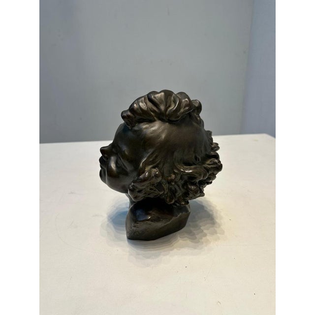 A finely crafted bronze bust depicting the head of a young child with detailed curls and a serene, upward gaze. This...