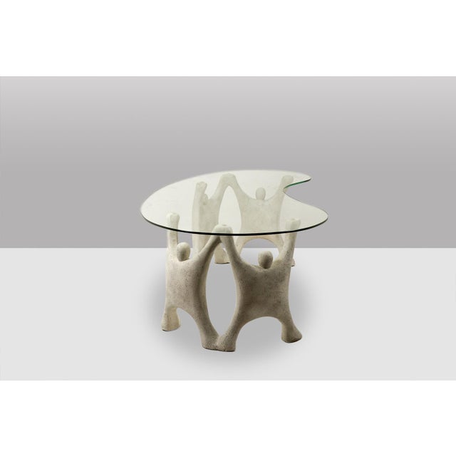 A Brutalist-style coffee table featuring a sculptural, organic base made of reconstituted stone and a glass top. from the...