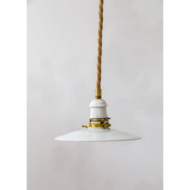A contemporary pendant light with a rope-covered cord, small milk glass saucer shade, and ceramic and brass canopy....