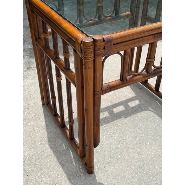A vintage 1970s end table crafted from bamboo and rattan, featuring an intricate open fretwork design and a clear glass...