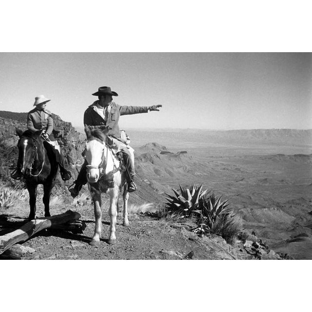 This black-and-white photograph captures two individuals on horseback in the vast expanse of the American Southwest. The...