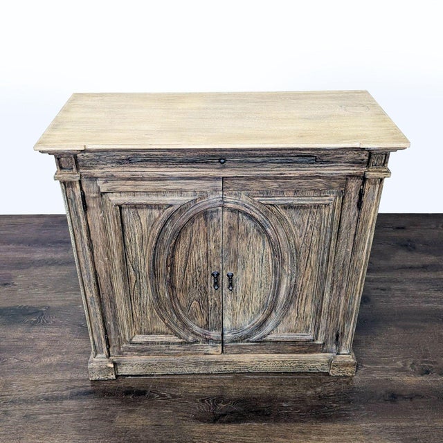 This wooden cabinet features a rustic finish and a unique oval design on the front doors. It includes a top tray surface...