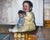 This oil painting on paper captures a joyful moment of a baby, seated on the floor, holding a doll while surrounded by...