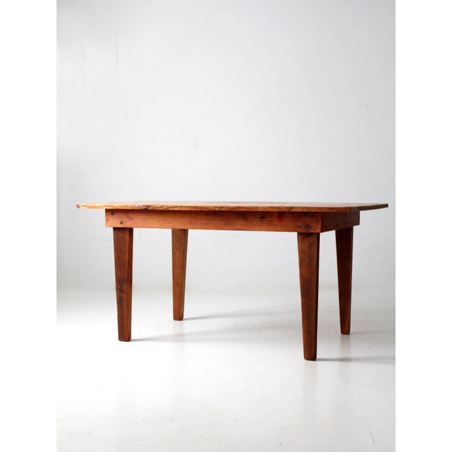 An antique primitive dining table featuring a wide plank top and simply tapered legs. The rustic wooden construction and...