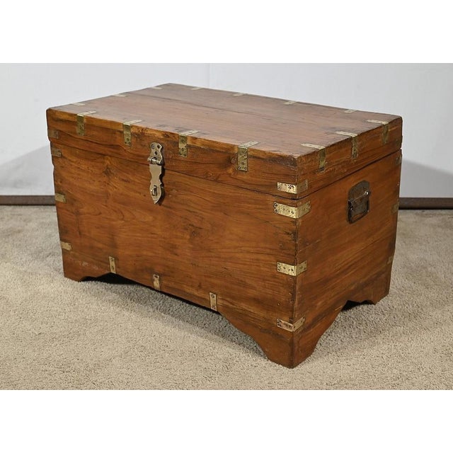 Wledge of solidity, the angles of this solid teak chest, are embellished with brass straps. The lid, mounted on two large...