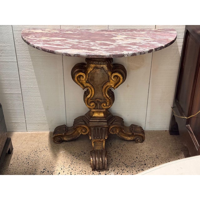 A pair of Italian console tables, each with a demilune top of rose and cream veined marble raised on a boldly carved...