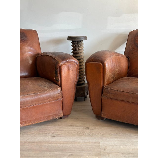 A pair of oversized French club chairs in original tan leather, deeply worn, with the rounded arms and low backs the form...