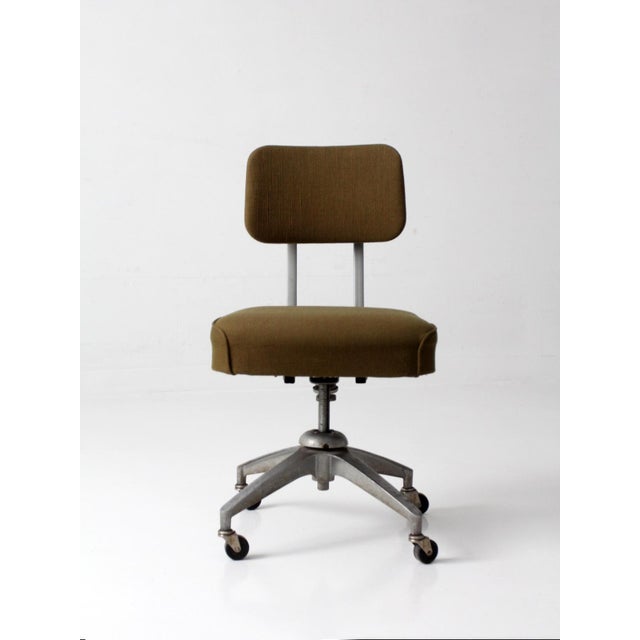 A mid-century industrial office chair featuring a clean, utilitarian silhouette and warm olive upholstery. The four-star...