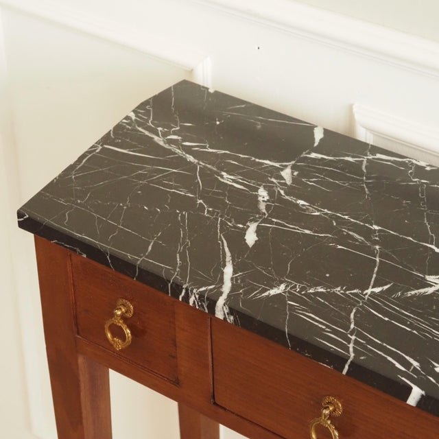 A narrow console table in the louis xvi style, likely made in the early 20th century, with a black marble top and tapered...