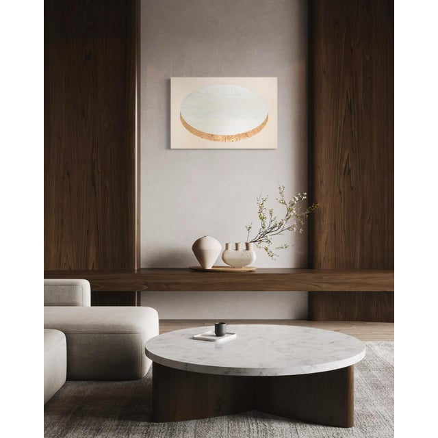 A soft, opalescent oval rests on warm taupe, its lower curve traced in a thin band of gold leaf that catches the light...