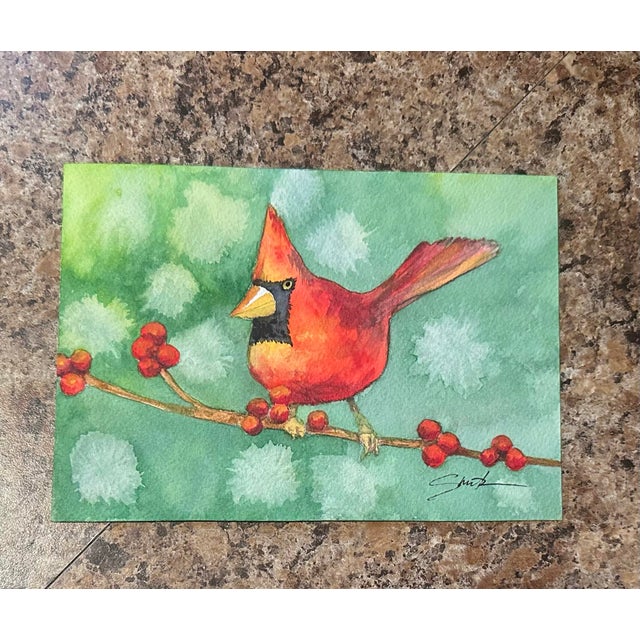 A vibrant red cardinal perches gracefully on a berry-laden branch, standing out against a soft, snowy green background....