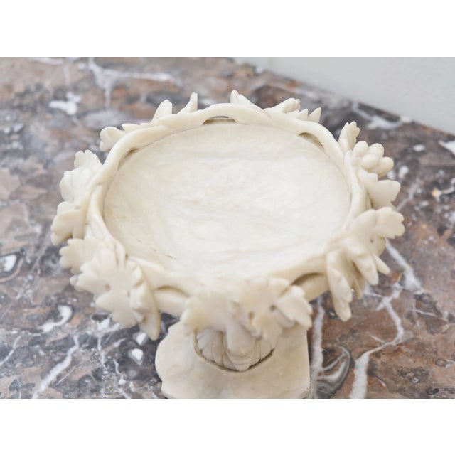 A decorative alabaster centerpiece featuring a finely carved rim adorned with leaf ornamentation. The central stem...