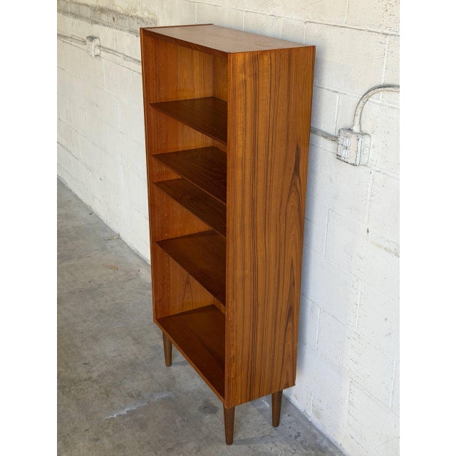 This tall teak bookcase by Poul Hundevad pairs a narrow profile with four open shelves and five display levels. Clean...