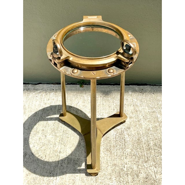 This vintage brass and glass porthole accent table channels nautical maritime design, its round glass top set securely...