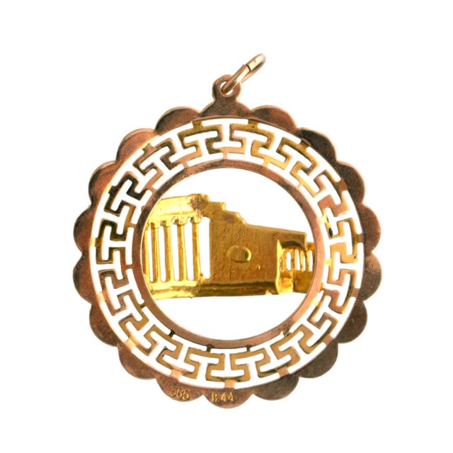 A circular charm. Crafted from 14k yellow gold, it features a Greek key pattern. Adorned with a detailed Parthenon design....