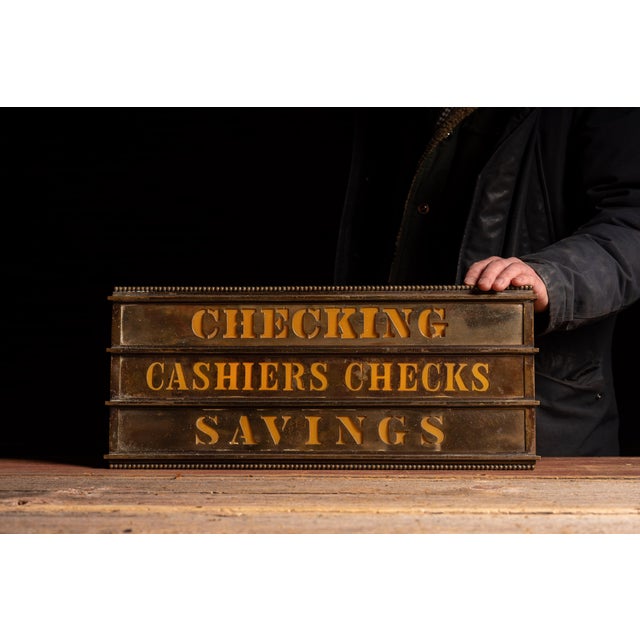 Illuminated brass teller sign with three stacked panels reading "CHECKING," "CASHIERS CHECKS," and "SAVINGS," each cut...