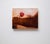 The artwork presents a serene composition featuring a hand resting on a textured surface, with a single red apple perched...