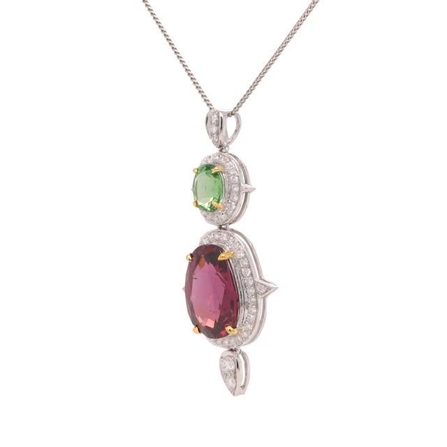 This platinum pendant showcases an oval tsavorite garnet above a larger pink‑red garnet, each encircled by a diamond halo....