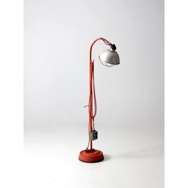 This is a vintage industrial floor lamp. The work lamp features an orange metal base and frame with an aluminum shade. It...