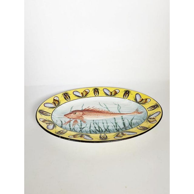 An elegant oval glazed ceramic platter, hand-painted with a fish design set against a stylized aquatic background. The rim...