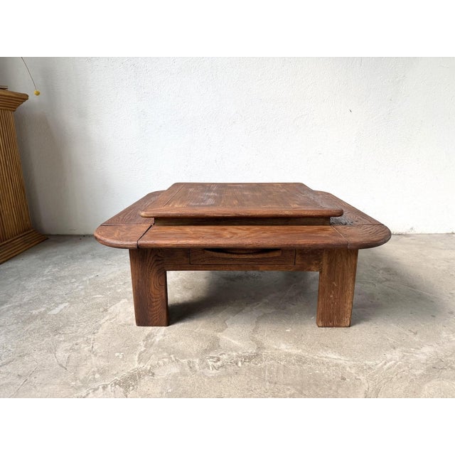 A tiered coffee table in solid oak, in a modernist and brutalist style, circa 1970. Its architectural lines are...