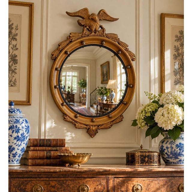 A Federal-style giltwood convex mirror crowned with a carved eagle, its wings outstretched above a laurel motif—an...