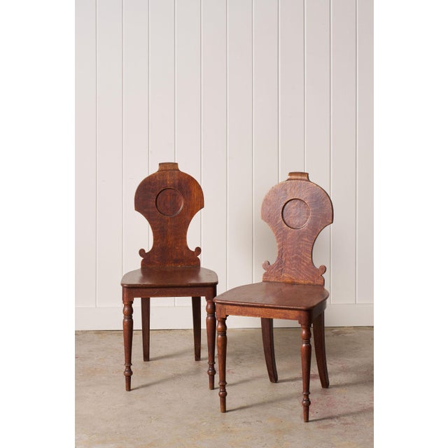 Pair of 19th-century oak hall chairs with carved backs, solid plank seats, and turned front stretchers. Constructed of oak...
