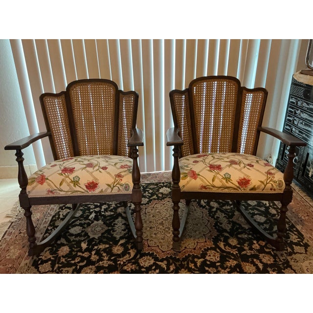 A charming pair of vintage rocking armchairs featuring woven cane backs, turned legs, and classic floral and bamboo...