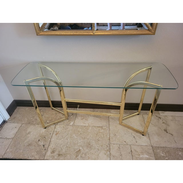 This contemporary console table features a clear rectangular glass top resting on a striking gold‑finished metal base with...