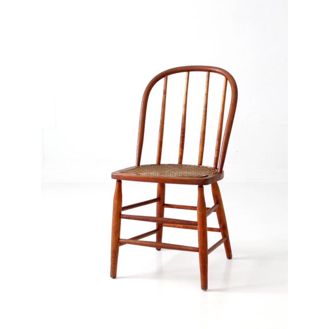 A graceful antique Windsor-style chair featuring a bowed spindle back, turned legs, and a beautifully hand-caned seat. Its...