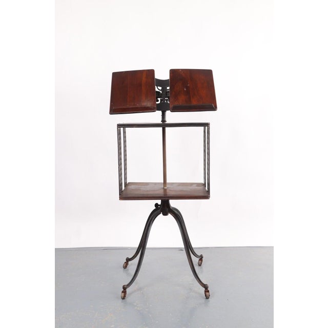 Victorian style dark toned wood library bookstand with a storage shelf on a four legged iron base with caster wheels. The...