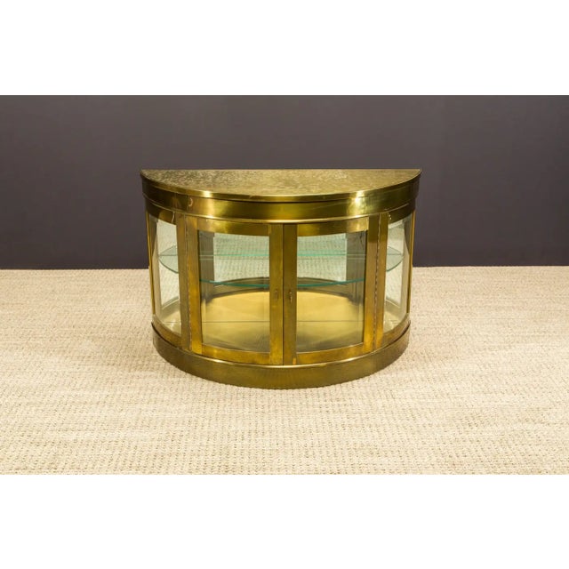 This incredible brass demilune display cabinet, produced by Mastercraft in the 1970s, features a mirrored and illuminated...