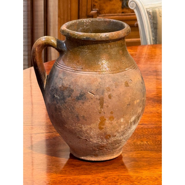 A small antique terra cotta jug with a rounded body, single pulled handle, and short flared neck. The surface shows a...