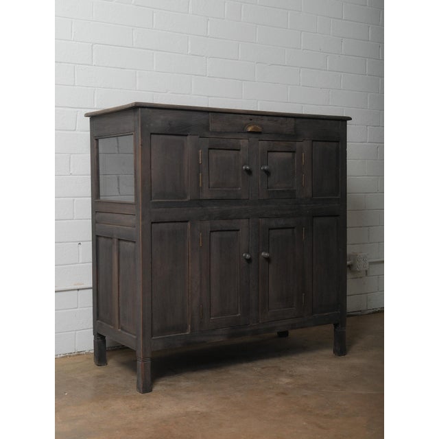A wooden sideboard with a dark, matte surface and a straightforward paneled construction. The upper section contains a...