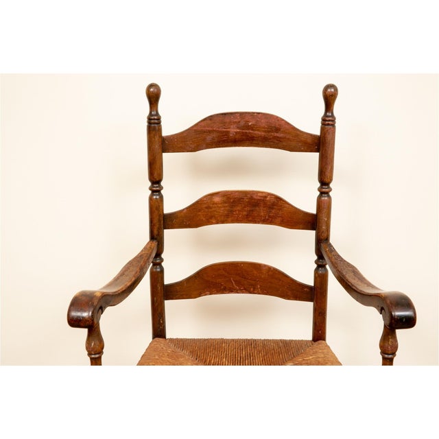 An antique fruitwood ladder-back armchair with a woven rush seat, tall curved slat-backs, turned finials, scrolled arms,...